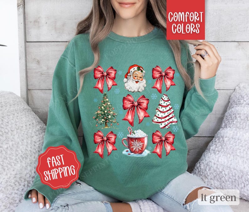 May include: A light green sweatshirt with a Christmas theme featuring Santa Claus, Christmas trees, bows, and a cup of hot chocolate. The sweatshirt has the text "Comfort Colors" on the top right corner and "Fast Shipping" on the bottom left corner.
