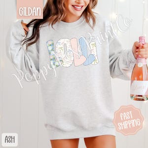 May include: A gray sweatshirt with the word "Lola" spelled out in colorful patchwork letters. The letters are made of different fabrics with floral, plaid, and gingham patterns.