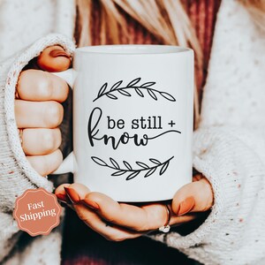 May include: White ceramic mug with black text and floral design that reads "be still + know".