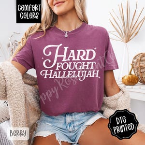 May include: Berry-colored Comfort Colors t-shirt with the words "Hard Fought Hallelujah" in white script. The shirt has a relaxed fit and is paired with denim shorts. The image also includes a DTG printed logo.