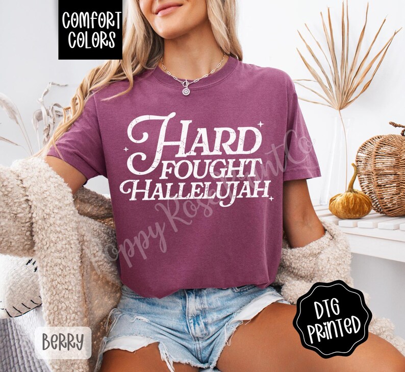 May include: Berry-colored Comfort Colors t-shirt with the words "Hard Fought Hallelujah" in white script. The shirt has a relaxed fit and is paired with denim shorts. The image also includes a DTG printed logo.