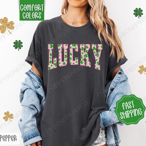 May include: A dark gray t-shirt with a pink "LUCKY" graphic. The letters are filled with green shamrock patterns.