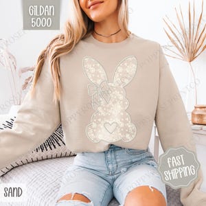 May include: A beige sweatshirt featuring a white lace bunny design. The bunny has a bow and a heart on its chest. The text "Gildan 5000" is visible in the top left corner of the image. The text "Sand" is visible in the bottom left corner of the image.