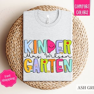 May include: Ash grey t-shirt with the text "Kinder Mrs. Wilson Garten" in colorful, playful letters. The letters are decorated with school-themed illustrations like pencils, apples, and a ruler.