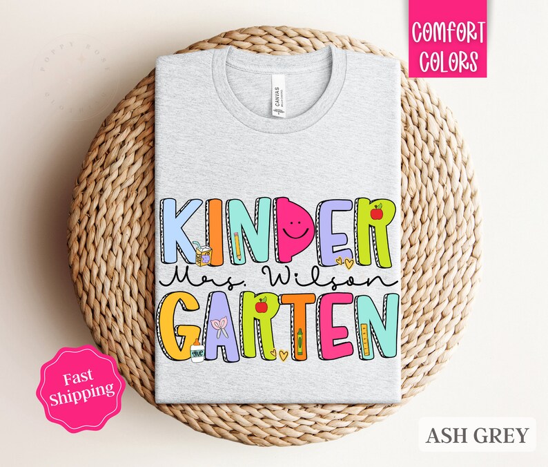 May include: Ash grey t-shirt with the text "Kinder Mrs. Wilson Garten" in colorful, playful letters. The letters are decorated with school-themed illustrations like pencils, apples, and a ruler.