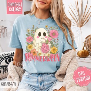 May include: Light blue t-shirt with a graphic of a ghost drinking a pink beverage, surrounded by flowers. The word "SUMMERWEEN" is printed in pink. The shirt also has the words "COMFORT COLORS", "CHAMBRAY", and "DTG PRINTED".
