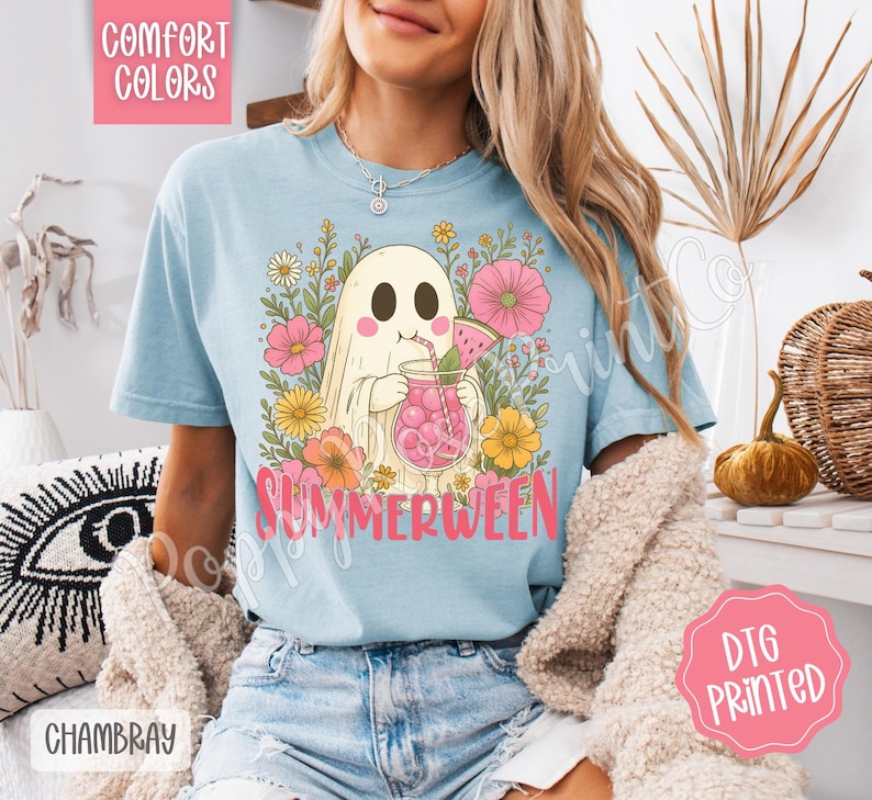 May include: Light blue t-shirt with a graphic of a ghost drinking a pink beverage, surrounded by flowers. The word "SUMMERWEEN" is printed in pink. The shirt also has the words "COMFORT COLORS", "CHAMBRAY", and "DTG PRINTED".