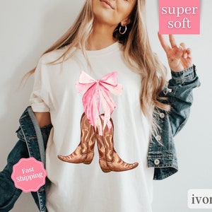 K&ouml;nnte beinhalten: Wei&szlig;es T-Shirt mit einer rosa Schleife und braunen Cowboy-Stiefeln. Der Text "super soft" befindet sich auf einem rosa Quadrat und der Text "ivory" auf einem wei&szlig;en Quadrat. Der Text "Fast Shipping" befindet sich auf einem rosa Kreis.