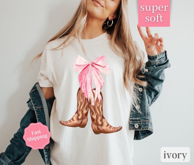 K&ouml;nnte beinhalten: Wei&szlig;es T-Shirt mit einer rosa Schleife und braunen Cowboy-Stiefeln. Der Text "super soft" befindet sich auf einem rosa Quadrat und der Text "ivory" auf einem wei&szlig;en Quadrat. Der Text "Fast Shipping" befindet sich auf einem rosa Kreis.
