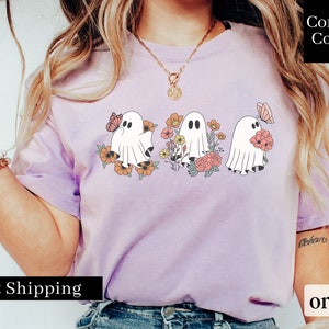Peut inclure: Un t-shirt violet avec un motif de trois fant&ocirc;mes tenant des fleurs et des papillons. Le texte "Comfort Colors" est dans le coin sup&eacute;rieur droit de l'image.
