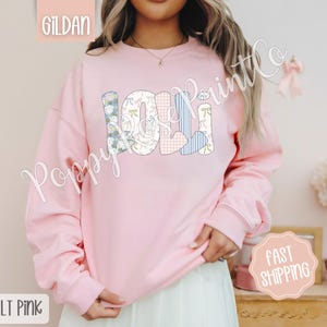 May include: A light pink sweatshirt with the word "LOLI" spelled out in colorful patchwork letters. The letters are decorated with floral and heart patterns.