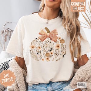 Pu&ograve; includere: T-shirt color avorio con un design floreale a forma di zucca e un fiocco. La zucca &egrave; piena di fiori, pipistrelli e zucche nei toni del pesca, marrone e crema. La maglietta ha le parole "Comfort Colors" e "DTG Printed".