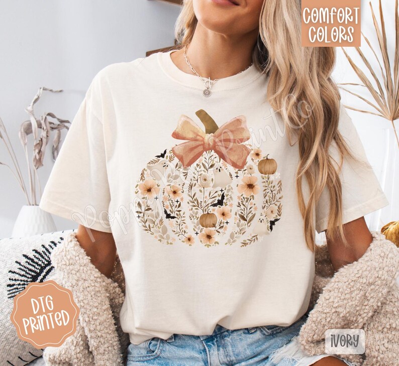 Pu&ograve; includere: T-shirt color avorio con un design floreale a forma di zucca e un fiocco. La zucca &egrave; piena di fiori, pipistrelli e zucche nei toni del pesca, marrone e crema. La maglietta ha le parole "Comfort Colors" e "DTG Printed".