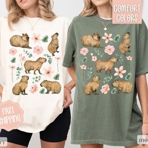 Pu&ograve; includere: Due t-shirt con un motivo di capibara e fiori. La maglietta a sinistra &egrave; avorio e la maglietta a destra &egrave; verde muschio. Il testo "COMFORT COLORS" &egrave; sulla maglietta a destra.