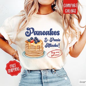 May include: A white t-shirt featuring a graphic of a stack of pancakes with blueberries and syrup, and the text "Pancakes & Panic Attacks! Buy one get one!" in a retro font.