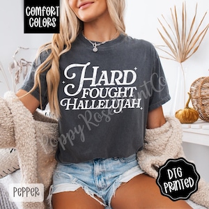 May include: Dark grey t-shirt with the words "Hard Fought Hallelujah" in a white, vintage-style font. The shirt is a Comfort Colors brand and is printed with DTG technology. The shirt is a Pepper color.