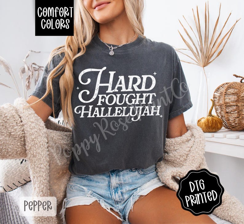 May include: Dark grey t-shirt with the words "Hard Fought Hallelujah" in a white, vintage-style font. The shirt is a Comfort Colors brand and is printed with DTG technology. The shirt is a Pepper color.