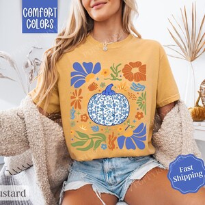 Op de afbeelding: Een mosterdgeel T-shirt met een blauw en wit pompoenontwerp. De pompoen is omringd door bloemen en bladeren in tinten oranje, geel en blauw. De tekst "Comfort Colors" is zichtbaar in de linkerbovenhoek van de afbeelding.