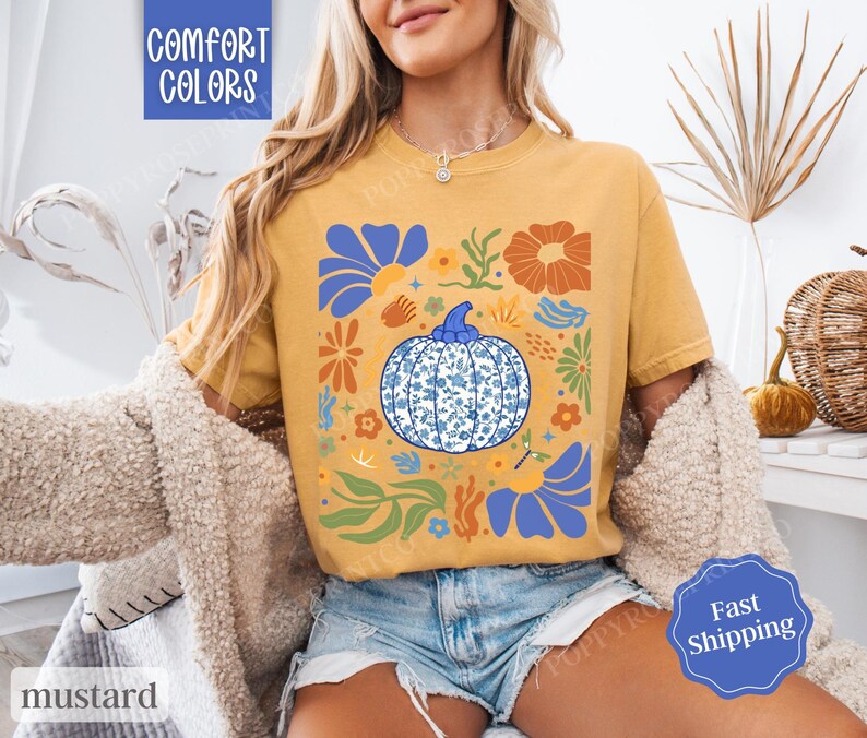Op de afbeelding: Een mosterdgeel T-shirt met een blauw en wit pompoenontwerp. De pompoen is omringd door bloemen en bladeren in tinten oranje, geel en blauw. De tekst "Comfort Colors" is zichtbaar in de linkerbovenhoek van de afbeelding.