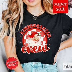 May include: Black t-shirt with a red and white checkered circle design featuring a cartoon Santa Claus and the text "Kindergarten Crew".