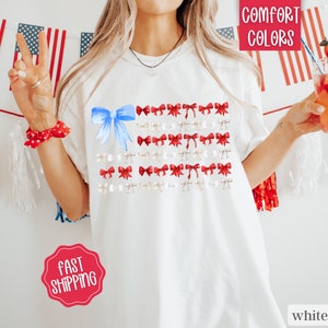 May include: White t-shirt featuring a design of the American flag made of bows. The flag design includes a large blue bow in the upper left corner, with red and white bows arranged in rows. The words "Comfort Colors" are in a red box in the upper right corner.