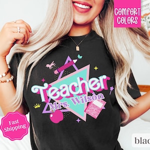 Puede incluir: Camiseta negra con un gráfico de estilo retro que dice "Teacher Mrs. Wilson". El gráfico presenta un triángulo rosa y azul, un lápiz, una corona, una manzana roja y un cuaderno.