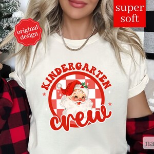 May include: White t-shirt with red text that says "Kindergarten Crew" and a red and white checkered circle with a cartoon Santa Claus inside.