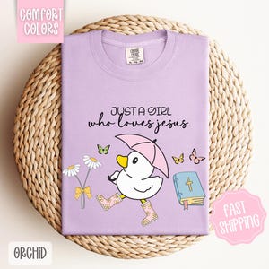 Puede incluir: Camiseta morada con un pato de dibujos animados que lleva botas de lluvia y sostiene un paraguas. El texto "Just a girl who loves Jesus" est&aacute; impreso en la camiseta. Hay una biblia y mariposas en la camiseta.