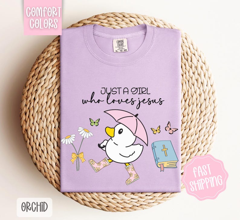 Puede incluir: Camiseta morada con un pato de dibujos animados que lleva botas de lluvia y sostiene un paraguas. El texto "Just a girl who loves Jesus" est&aacute; impreso en la camiseta. Hay una biblia y mariposas en la camiseta.