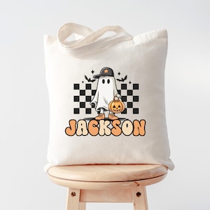 K&ouml;nnte beinhalten: Eine wei&szlig;e Stofftasche mit einem schwarz-wei&szlig;en Karomuster, einem Cartoon-Geist, der einen Hut tr&auml;gt und einen K&uuml;rbis h&auml;lt, und dem Text "JACKSON".