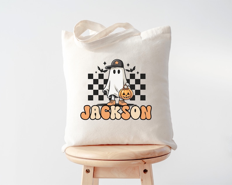 K&ouml;nnte beinhalten: Eine wei&szlig;e Stofftasche mit einem schwarz-wei&szlig;en Karomuster, einem Cartoon-Geist, der einen Hut tr&auml;gt und einen K&uuml;rbis h&auml;lt, und dem Text "JACKSON".