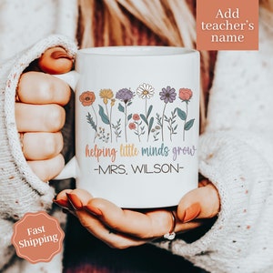 Może przedstawiać: Biały ceramiczny kubek z kolorowym kwiatowym wzorem i napisem "helping little minds grow" oraz imieniem nauczyciela. Kubek ma uchwyt i jest trzymany przez osobę.