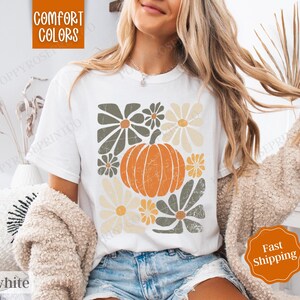 Puede incluir: Camiseta blanca con un dise&ntilde;o gr&aacute;fico de una calabaza y flores en tonos verdes, amarillos y naranjas. El texto "Comfort Colors" es visible en la esquina superior izquierda de la imagen.