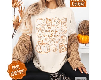 May include: Cream-colored t-shirt with a fall-themed graphic that includes a pumpkin, bow, leaf, and the words "cozy vibes." The shirt is paired with black pants and a brown cardigan. The words "Comfort Colors" are in the top right corner.