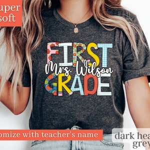 May include: Dark heather grey t-shirt with the text "First Grade" in colourful letters with a pattern of apples and school supplies. The teacher's name is customisable and is shown as "Mrs. Wilson" in this image.