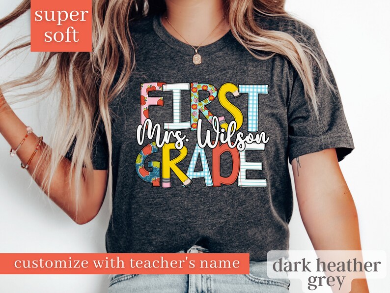 May include: Dark heather grey t-shirt with the text "First Grade" in colourful letters with a pattern of apples and school supplies. The teacher's name is customisable and is shown as "Mrs. Wilson" in this image.