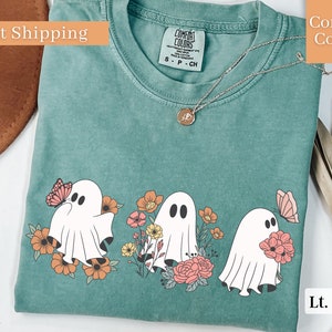 Peut inclure: Un t-shirt vert clair avec un motif de trois fant&ocirc;mes tenant des fleurs et des papillons. Les fant&ocirc;mes sont blancs avec des yeux et une bouche noirs. Les fleurs sont roses, orange et jaunes. Les papillons sont roses et orange.