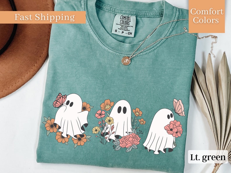 Peut inclure: Un t-shirt vert clair avec un motif de trois fant&ocirc;mes tenant des fleurs et des papillons. Les fant&ocirc;mes sont blancs avec des yeux et une bouche noirs. Les fleurs sont roses, orange et jaunes. Les papillons sont roses et orange.