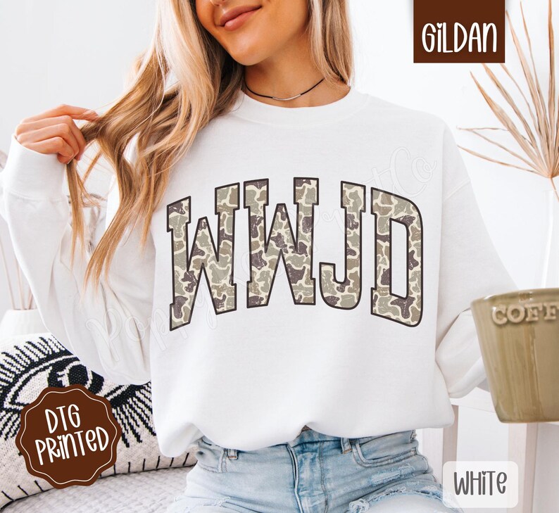 May include: White sweatshirt with the letters WWJD in a camouflage pattern across the chest. The sweatshirt is a Gildan brand and has a crew neck. The words "DTG Printed" are on a brown sticker.