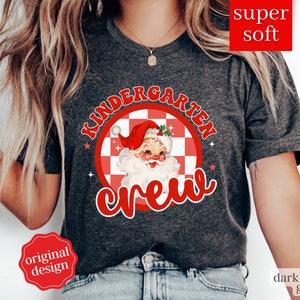 May include: Dark heather grey t-shirt with a red and white checkered circle design featuring a vintage Santa Claus illustration and the text "Kindergarten Crew".