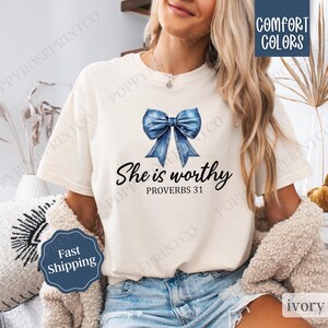 May include: A white t-shirt with a blue denim bow and the text "She is worthy Proverbs 31" printed on it.