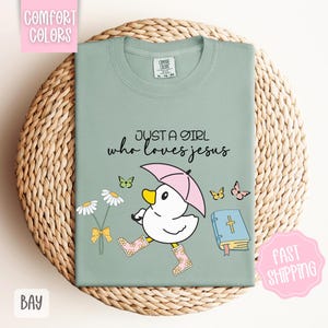Puede incluir: Una camiseta verde claro con el texto "Just a girl who loves Jesus" y una ilustraci&oacute;n de dibujos animados de un pato con botas de lluvia y sosteniendo un paraguas. El pato camina junto a un libro con una cruz y flores.