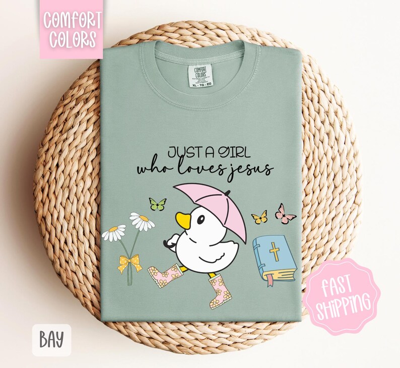 Puede incluir: Una camiseta verde claro con el texto "Just a girl who loves Jesus" y una ilustraci&oacute;n de dibujos animados de un pato con botas de lluvia y sosteniendo un paraguas. El pato camina junto a un libro con una cruz y flores.