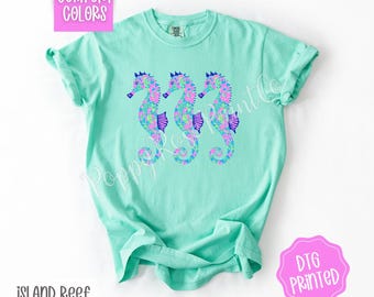 May include: A turquoise t-shirt with a design of three seahorses with floral patterns in shades of pink, blue, and purple. The text "Island Reef" is printed below the design. The text "DTG Printed" is printed on a pink circle in the lower right corner.