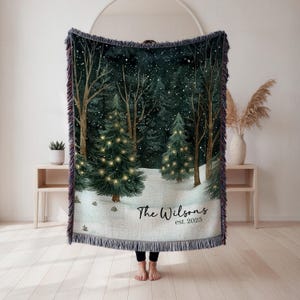 May include: A woven Christmas throw blanket featuring a winter forest scene with snow-covered ground, trees, and twinkling lights. The blanket has a fringed edge and the text "The Wilsons est. 2025". The color palette includes green, white, and brown.