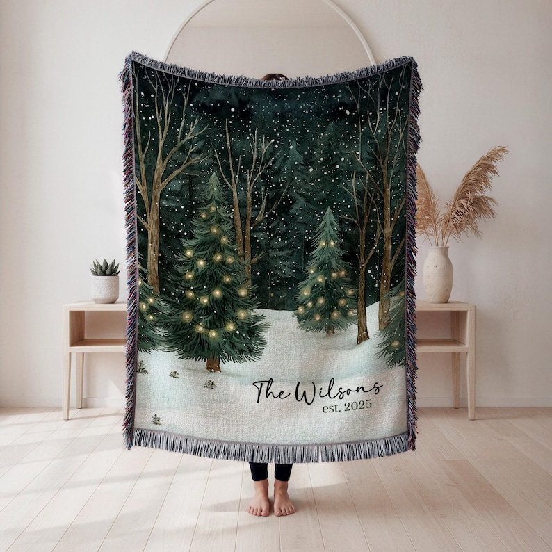 May include: A woven Christmas throw blanket featuring a winter forest scene with snow-covered ground, trees, and twinkling lights. The blanket has a fringed edge and the text "The Wilsons est. 2025". The color palette includes green, white, and brown.
