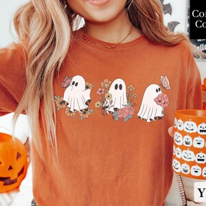 Peut inclure: Un t-shirt orange br&ucirc;l&eacute; avec un motif de trois fant&ocirc;mes tenant des fleurs et des papillons. Le texte "Comfort Colors" est visible en arri&egrave;re-plan.