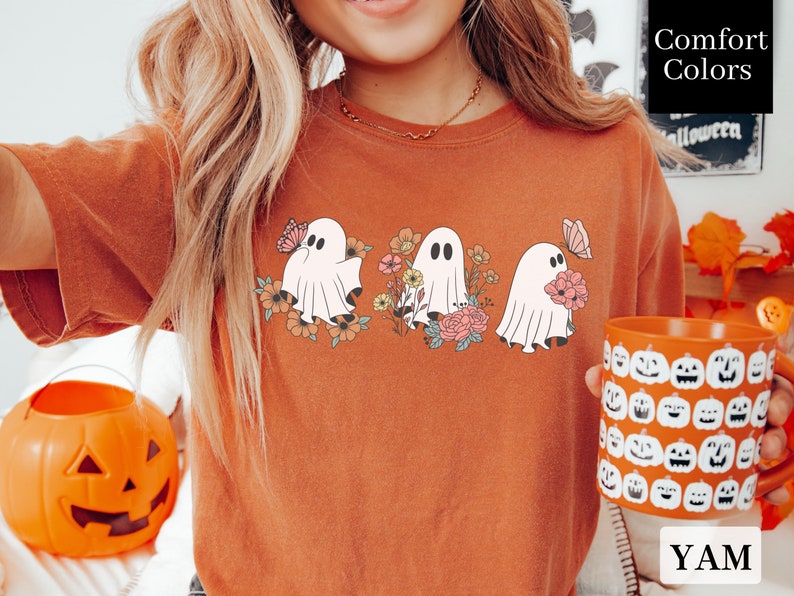 Peut inclure: Un t-shirt orange br&ucirc;l&eacute; avec un motif de trois fant&ocirc;mes tenant des fleurs et des papillons. Le texte "Comfort Colors" est visible en arri&egrave;re-plan.