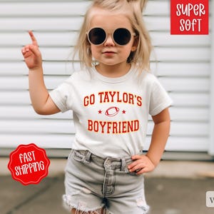 May include: White t-shirt with red and yellow text that says "Go Taylor's Boyfriend" with a football graphic. The shirt is on a young girl wearing sunglasses and denim shorts.