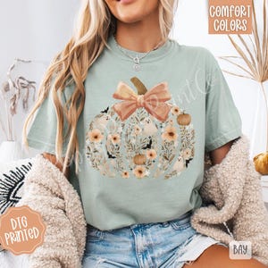 Pu&ograve; includere: T-shirt verde chiaro con un design a zucca pieno di fiori, pipistrelli e fantasmi. La zucca ha un grande fiocco color pesca e un gambo marrone. Le parole "Comfort Colors" sono stampate in alto a destra.